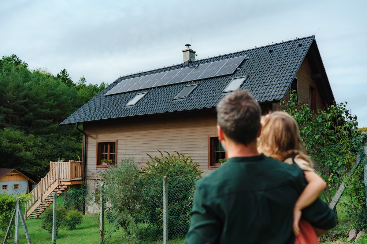 Rear view of dad holding her little girl in arms and looking at their house with solar panels