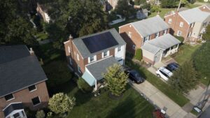 Aerial view of a residential neighborhood with brick houses. The main house boasts solar panels on the roof, a driveway with a parked car, and trees surrounding the property, showcasing modern roofing solutions.