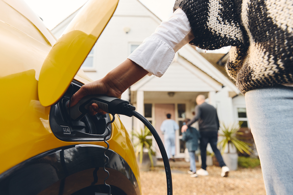 A person is installing an at-home EV charger and plugging it into a yellow electric vehicle in front of a house, while three people walk towards the entrance.