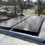 Solar panels installed on a flat rooftop capture solar energy, surrounded by bare trees and overlooking a residential street.