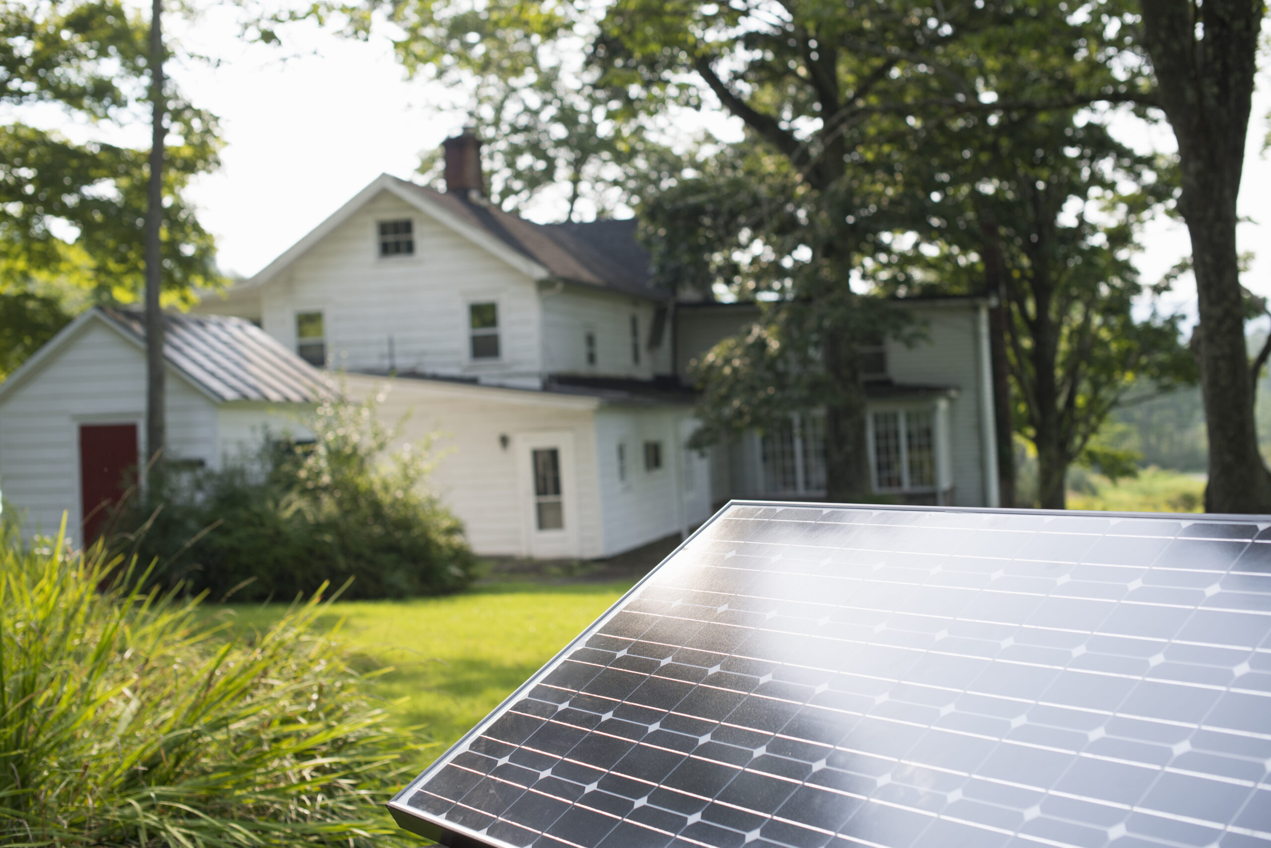 A solar panel is installed in the yard of a two-story white house surrounded by greenery and trees, taking advantage of Pennsylvania's government incentives to promote renewable energy.